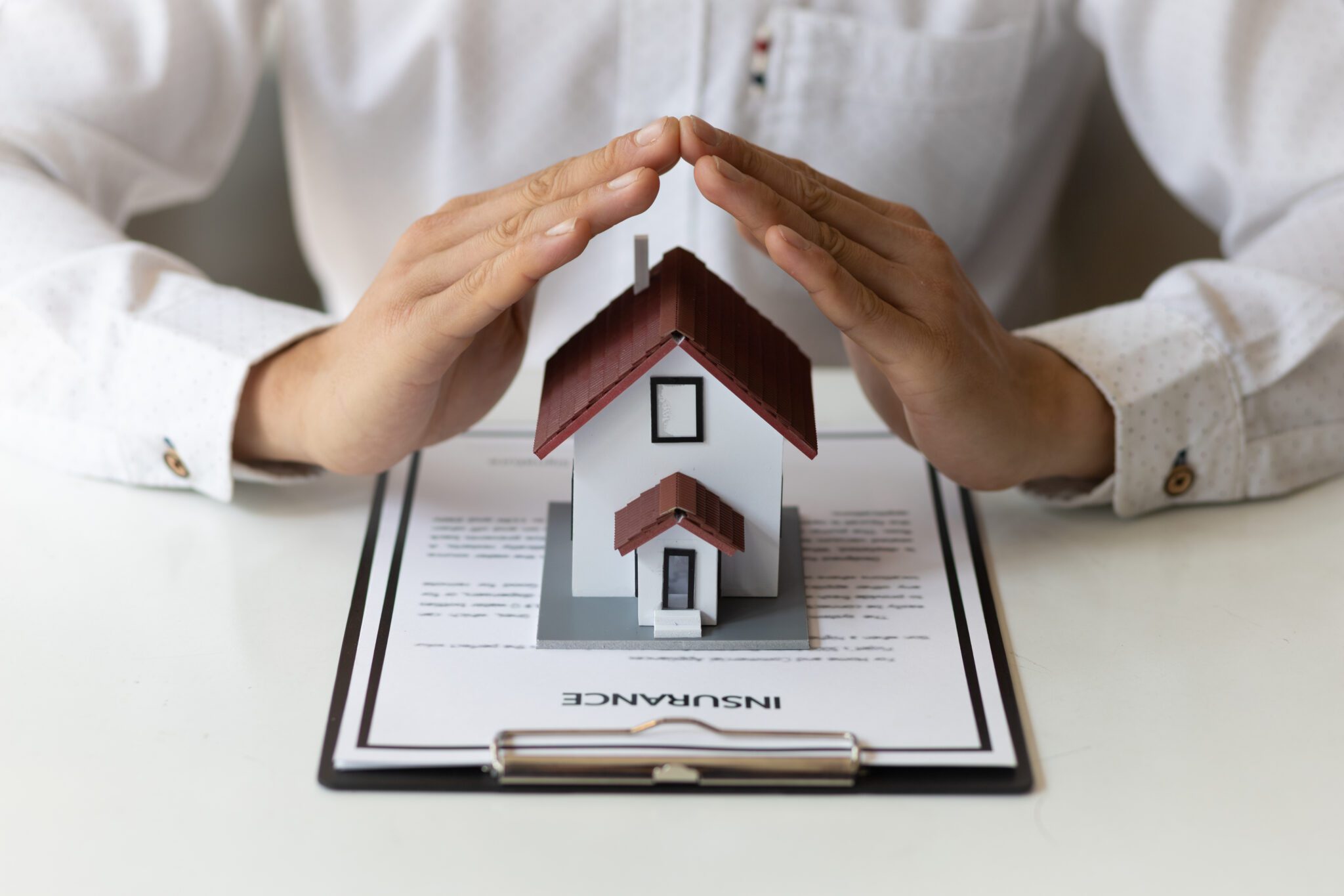 Hands protect a small house model on top of an insurance document.