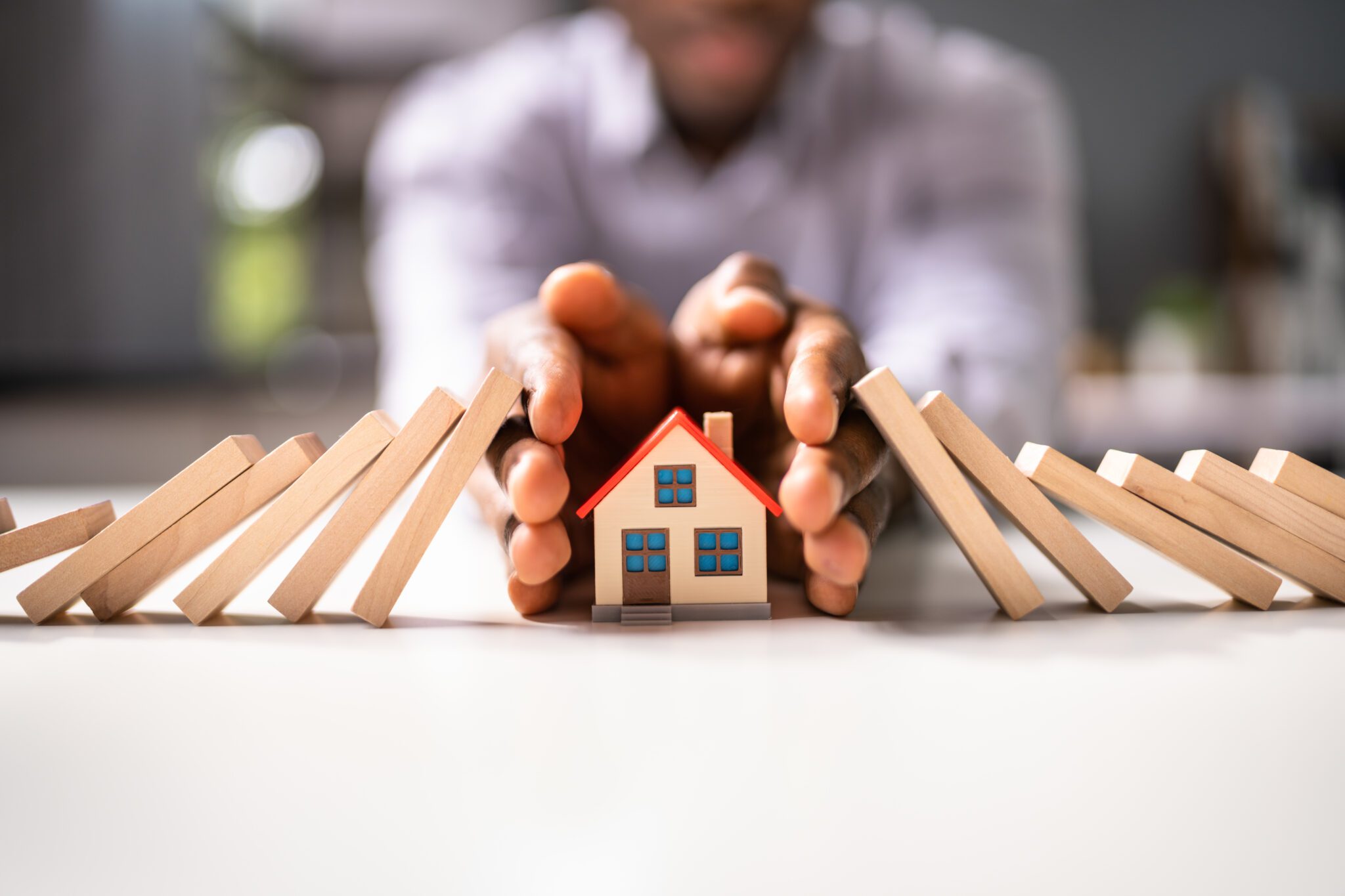 Hands protecting a small house model from falling dominoes.